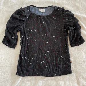 Svart blus med blommigt mönster från Holly & Whyte - Säljer en svart blus från Holly & Whyte med små blommiga detaljer i grönt, rött och vitt. Blusen har trekvartsärm med puff och är tillverkad i ett lätt, syntetiskt material. Perfekt för dig som gillar färg och mönster!