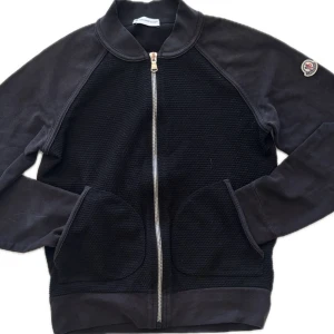 Mörkblå kofta från Moncler - Moncler kofta, Snygg mörkblå zipjacka från Moncler med dragkedja framtill och två fickor. Jackan har lång ärm och klassisk Moncler-logga på ärmen. Den har ända sen defekt som kan ses på bild 3(går att tvätta bort). Perfekt för en stilren och sportig look. Priset kan ändras i snabb affär