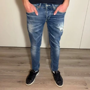 Blå jeansbyxor från Dondup - Säljer ett par blå jeansbyxor från Dondup i klassisk tvätt med slitningar och lätt trasiga detaljer på låret. Byxorna har normal passform, raka ben och femficksdesign. Midjan är midwaist och materialet är bomull med lite stretch.