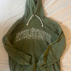 Olivgrön hoodie med tryck från HM - Säljer en olivgrön hoodie från HM med vit text på bröstet och justerbar huva med vita snören. Perfekt för en avslappnad stil och sköna dagar. Mjuk insida och klassisk känguruficka framtill.