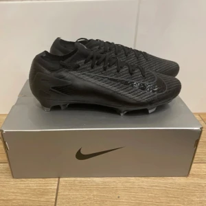 Nike Zoom Vapor 16 Elite FG fotbollsskor - Säljer ett par helsvarta Nike Zoom Vapor 16 Elite FG fotbollsskor. Skorna har en modern, strömlinjeformad design med snörning och en lätt ovandel i syntetmaterial. Yttersulan är anpassad för spel på naturgräs och har en blå detalj undertill.