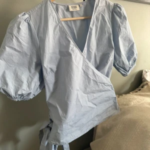 Ljusblå omlottblus från Magasin du Nord - Säljer en ljusblå omlottblus från Magasin du Nord i ekologisk bomull. Blusen har puffiga korta ärmar, v-ringning och knyt i sidan. Perfekt för vår och sommar!