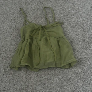 Grön volangig linnetopp - Säljer en söt grön linnetopp med tunna axelband och volangig nederdel. Toppen har knytning framtill och är perfekt för varma dagar. Lätt och luftig modell som ger en avslappnad känsla.