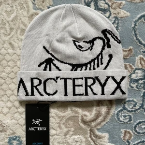 Vit mössa från Arc'teryx med logga - Snygg vit mössa från Arc'teryx med svart logotyp och grafiskt skelettmönster framtill. Mössan är stickad i mjukt material och har uppvikt kant. Perfekt för kalla dagar när du vill ha både stil och värme.