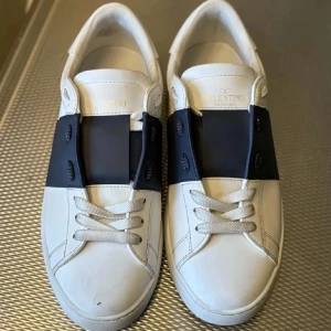 Valentino Garavani vit/svart - Snygga Valentino Garavani Open sneakers i vitt skinn med bred svart rem över vristen och coola nitar baktill. Klassisk rund tå, platt sula och vita. Några defekter på skorna sen ja la upp bilden så fråga så kan jag visa