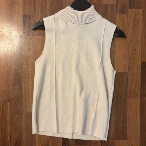 Vit ärmlös polotopp - Snygg vit polotopp utan ärmar med ribbad polo krage. Perfekt att styla med jeans eller kjol för en clean look. Mjuk och stickad känsla i materialet, passar både till lager-på-lager eller som den är.