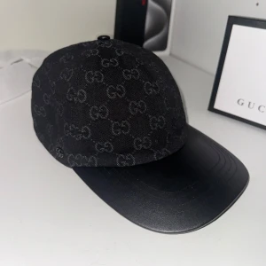 Svart keps från Gucci med GG-mönster - Snygg svart keps från Gucci med klassiskt GG-monogram över hela kepsen. Skärmen är i svart skinn och baksidan har justerbart läderband med präglad Gucci-logga. Kepsen är tillverkad i bomull och läder, perfekt för dig som vill ha en exklusiv streetstyle-look.