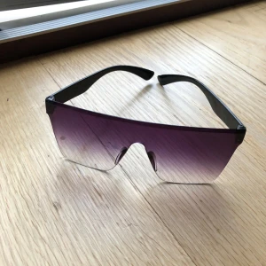 Svarta shield-solglasögon med lila ton - Säljer ett par futuristiska shield-solglasögon med svarta skalmar och stora, lila tonade linser som går över hela ansiktet. Glasögonen har en cool, sportig vibe och passar perfekt för dig som vill sticka ut med en edgy stil.
