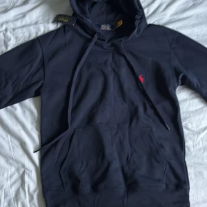 Mörkblå hoodie från Polo Ralph Lauren - Snygg mörkblå hoodie från Polo Ralph Lauren med klassisk röd broderad logga på bröstet. Tröjan har huva med snörning och en stor magficka. Tillverkad i mjuk bomull och har långa ärmar. Perfekt för en chill och stilren look.
