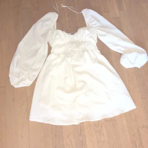 Vit kort klänning från Bershka - Superfin vit kort klänning från Bershka med puffiga långärmar och söt rynkad detalj vid midjan. Klänningen har en lätt struktur i tyget och knytband upptill. Perfekt för dig som gillar romantisk och trendig stil.