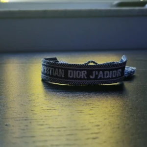 Christian Dior J'Adior armband blå - Snyggt vävt armband från Christian Dior med texten 'J'Adior' i vitt på blå bakgrund. Armbandet har justerbar knytning med tofsar i ändarna och ger en cool och trendig vibe. Perfekt accessoar för att lyfta din outfit.