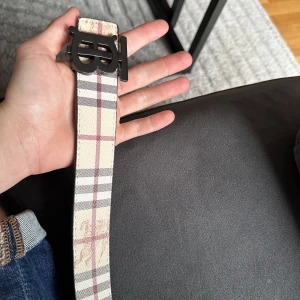 Burberry rutig skärp med TB-spänne - Säljer ett snyggt Burberry-bälte med klassiskt rutigt mönster i beige, svart, grått och rött. Bältet har ett stort TB-monogram i metall som spänne och är gjort i skinn. Insidan är svart med rutigt mönster. Perfekt accessoar för att lyfta din outfit.