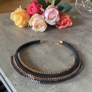 Svart och guldigt statement-halsband - Snyggt statement-halsband med svart flätad textil och detaljer i guld och mörkgrå metall. Halsbandet har flera lager med kedjor och fyrkantiga länkar som ger en cool och modern look. Justerbar kedja i nacken för perfekt passform.