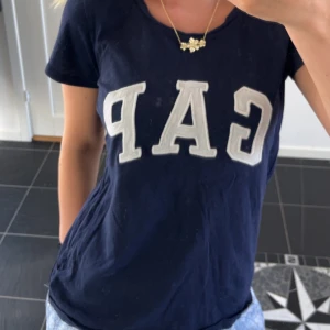 Mörkblå GAP t-shirt med tryck - Snygg mörkblå t-shirt från GAP med stort vitt logotryck över bröstet. Klassisk rund halsringning och korta ärmar. Perfekt för en avslappnad och cool stil.