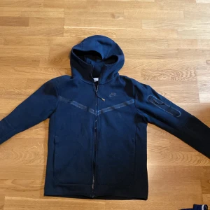 Mörkblå hoodiejacka från Nike - Snygg svart hoodiejacka från Nike med hel dragkedja och huva. Jackan har en diskret Nike-logga på bröstet, en praktisk ficka med dragkedja på ärmen och coola detaljer i form av svarta band över bröstet. Perfekt för dig som gillar sportig och stilren look. Inga skador och den har använts cirka 2 gånger. Är S men passar också M 