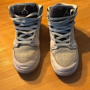 Nike Air Jordan 1 Mid ICE blue  - Säljer ett par Nike Air Jordan 1 Mid i vitt och svart skinn med klassisk svart swoosh och Jordan-logga på sidan. Skorna har perforerad tå, högt skaft och ljusgrå snörning. Perfekta för dig som gillar streetstyle och vill sticka ut med en ikonisk sneaker.