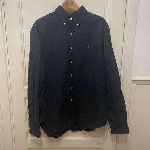 Svart skjorta från Ralph Lauren - Klassisk svart skjorta från Ralph Lauren med button-down krage och broderad logga på bröstet. Skjortan har lång ärm och vita knappar framtill. Perfekt till dig som gillar stilrena plagg med tidlös design.