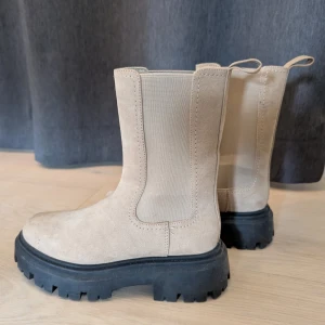 Beiga chunky chelsea boots i mocka - Snygga beiga chelsea boots i mocka med grov svart sula och rund tå. Boots med elastiska paneler på sidorna och dragflikar bak för enkel påtagning. Perfekta för dig som gillar en trendig och robust look.