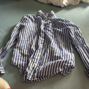 Blåvit randig skjorta från H&M - Snygg blåvit randig skjorta från H&M i storlek 152. Skjortan har klassisk krage, långa ärmar och knäppning framtill. Tillverkad i mjuk bomull som känns skön mot huden. Perfekt för dig som gillar en stilren och fräsch look.