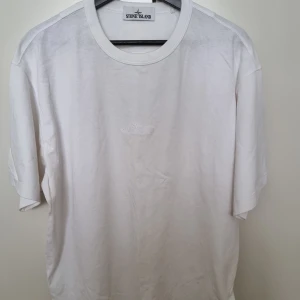 Vit t-shirt från Stone Island - Vit t-shirt från Stone Island med broderad logga framtill. Klassisk rund halsringning och korta ärmar. Tillverkad i mjuk bomull som känns skön mot huden. Enkel och stilren design som passar perfekt till jeans eller shorts.