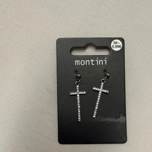 Glittriga korsörhängen från Montini - Snygga örhängen från Montini med korsform, täckta av glittrande stenar. De är silverfärgade och hänger snyggt från örat, perfekta för att ge din look lite extra edge. Enkla att matcha med olika outfits och riktigt trendiga.