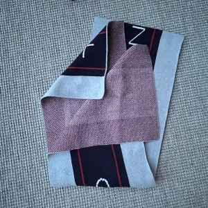 Stickad halsduk från GANT i grått och rött - Snygg stickad halsduk från GANT med breda grå partier, svart mitt med röda ränder och vit GANT-logga. Ena sidan har ett tätt rödvitt mönster. Perfekt accessoar för kalla dagar och riktigt stilren look. Inte använd.