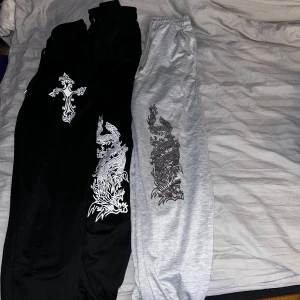 Svarta och grå mjukisbyxor med draktryck - Tre par mjukisbyxor, två svart och ett grått, båda med coolt draktryck längs benet. Det andra svarta paret har ett stort vitt kors. Byxorna har resår i midjan och är perfekta för en avslappnad streetstyle.