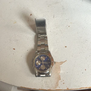 Blå och silverfärgad klocka Marble - Stilren klocka från Marble med blå urtavla och tre silvriga subdials. Boetten och länken är i blankt silverfärgat metall, vilket ger en klassisk och clean look. Perfekt accessoar för dig som gillar detaljer och snygg design.