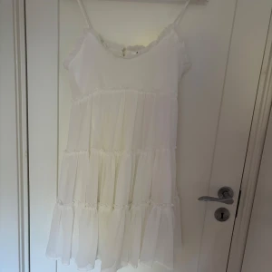 Vit volangklänning från DM Retro - Superfin vit kortklänning från DM Retro med tunna axelband, volanger och knappar i ryggen. Klänningen har flera lager med rynkade volanger och en luftig känsla, perfekt för varma dagar. Söt och romantisk stil som passar till sommarens alla tillfällen.