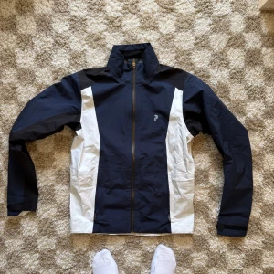 Peak Performance Gore-Tex vindjacka - Snygg vindjacka från Peak Performance i marinblått och vitt med svarta detaljer. Jackan har hel dragkedja framtill, hög krage och sportig design. Perfekt på golfbanan eller till hiking nu på sommaren. Superfint skick.