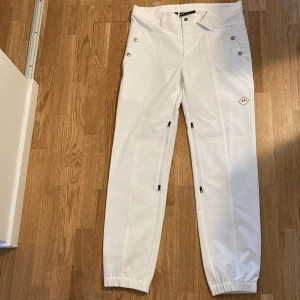 Vita täckbyxor från J.Lindeberg - Snygga vita täckbyxor från J.Lindeberg med dragkedjor vid benen och elastiska benslut. Byxorna har knappar i midjan, bälteshällor och en stilren look. Perfekta för vintern och outdoor-aktiviteter.