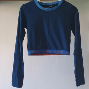 Blå croppad träningströja Only Play - Snygg croppad långärmad träningströja från Only Play i marinblått med ljusblå kant vid halsen. Nedtill finns ett elastiskt band med texten WORKOUT i orange. Perfekt för gymmet eller sportiga outfits.