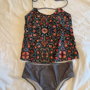 Blommig bikini med grå underdel - Helt ny, snygg bikini med blommigt mönstrat linne i mörkblått, rött och orange samt grå, enkel bikiniunderdel. Överdelen har smala axelband och färgstarka blommor som ger en unik look. Perfekt för stranden eller poolen. Enbart testad.