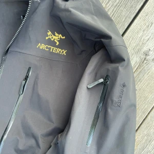 Arcteryx jacka - Svart vindjacka från Arc'teryx med GORE-TEX Pro-material, perfekt för dig som vill ha något riktigt tåligt. Jackan har huva, flera dragkedjeförsedda fickor och snygg gul logga på bröstet. Funktionell och stilren design med justerbara detaljer.