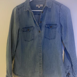 Ljusblå jeansskjorta Hampton Republic - Ljusblå jeansskjorta från Hampton Republic med klassisk krage, två bröstfickor med knappar och långa ärmar. Skjortan har snygga metallknappar och en avslappnad passform. Perfekt att styla lager på lager eller bära som den är.