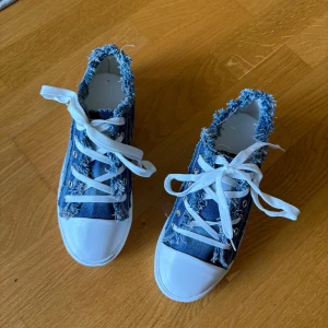 Blå denim sneakers med fransad kant - Säljer ett par unika liknande converse i blå denim med fransade kanter och vita snören. Skorna har vit gummisula och tå, samt coola detaljer som ger en avslappnad streetstyle-vibe. Perfekta för dig som gillar en trendig och avslappnad look.