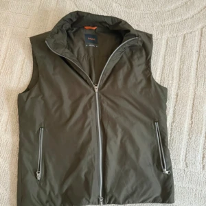 Olivgrön dunväst från Swims - Snygg olivgrön dunväst från Swims med hög krage och tvåvägsdragkedja framtill. Västen har två fickor med dragkedja och en stilren, minimalistisk look. Perfekt att slänga över en hoodie eller tröja för extra värme. Materialet är lätt och smidigt.