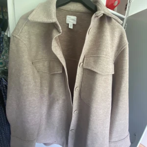Beige overshirt med fickor XS - Snygg beige overshirt från H&M i storlek XS. Jackan har klassisk krage, stora bröstfickor med lock och knäppning framtill. Perfekt lager-på-lager-plagg med avslappnad passform och lång ärm. Enkel att styla till både jeans och kjol.