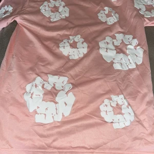 Rosa shorts och t-shirt från Denim Tears  - Säljer ett par rosa shorts från Denim Tears med vita blommiga tryck över hela plagget. Shortsen har elastisk midja med snörning för skön passform. Perfekta för en chill sommarlook och sticker ut med sitt unika mönster. 