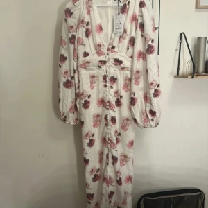 Blommig långklänning från Gina Tricot - Superfin långklänning med ballongärmar från Gina Tricot. Klänningen är vit med rosa och vinröda blommor, har djup v-ringning och knappar framtill. Perfekt för dig som vill sticka ut med romantiska vibes och snygg passform. Slutsåld överallt