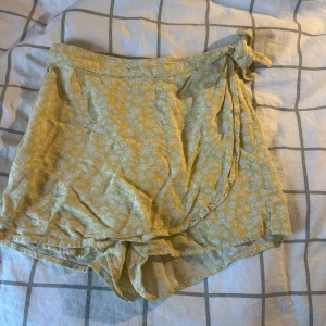 Gul kjol - Snygg gul kjol från Kappahl med vitt blommigt mönster och inbyggda shorts. De har en knytning i sidan och är tillverkade i ett lätt och luftigt material, perfekta för varma dagar. Passformen är normal och shortsen har en somrig vibe. Storleken är 140 men passar modellen som har stl 146!