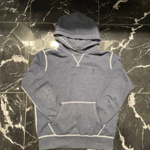 Blå hoodie från Polo Ralph Lauren - Snygg blå hoodie från Polo Ralph Lauren med klassisk broderad logga på bröstet. Tröjan har huva, magficka och kontrastsömmar i vitt. Perfekt för dig som gillar en avslappnad och stilren look.