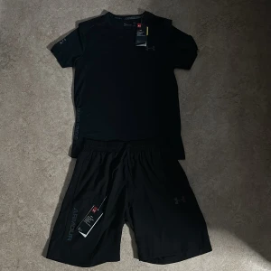 Svart Under Armour set - Svart set från Under Armour i sportig design med korta ärmar och färgglada logodetaljer på bröstet och sidan. Tillverkad i ett lätt och snabbtorkande funktionsmaterial som passar perfekt till träning. Snygg passform och diskreta tryck för en clean look.