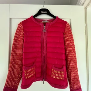 Rosa dunjacka från Moncler - Snygg rosa dunjacka från Moncler med stickade ärmar i rosa, orange och gult. Jackan har quiltad front, två fickor med stickade detaljer och dragkedja med Moncler-logga. Perfekt statement-plagg för dig som vill sticka ut.