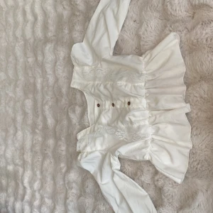 Vit blus med spets och knappar - Supersöt vit blus med v-ringning, bruna knappar framtill och volang nertill. Blusen har långa ärmar med lätt puff och breda manschetter. Spetsdetaljer längs bröstet ger en romantisk vibe. Perfekt för dig som gillar vintageinspirerat och feminina detaljer.