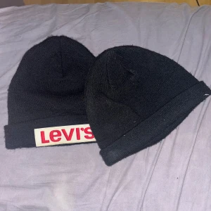 Svarta Mössor - Två svarta mössor i klassisk modell. Ena mössan har en tydlig Levi's-logga i rött och vitt framtill, medan den andra (Är inte Levi’s) är helt utan tryck. 🌹