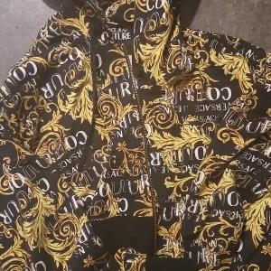 Versace Jeans Couture hoodie med guldmönster - Säljer en svart hoodie från Versace Jeans Couture med ikoniskt guldigt barockmönster och stora logotexter. Tröjan har huva, dragkedja och långa ärmar. Perfekt statement-plagg för dig som gillar att sticka ut. Nypriss 5899
