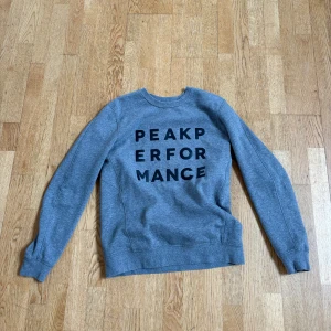 Grå sweatshirt från Peak Performance - Snygg grå sweatshirt från Peak Performance med svart tryck på bröstet. Klassisk rund halsringning och långa ärmar. Perfekt för dig som gillar en enkel och stilren look. Mjuk insida och ribbade muddar för extra komfort.