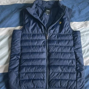 Väst från lyle & Scott  - Snygg marinblå dunväst från Lyle & Scott Junior med gul logga på bröstet. Västen är quiltad med horisontella sömmar och har dragkedja framtill. Perfekt att slänga över en hoodie för en chill look.
