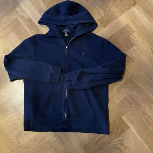 Mörkblå hoodie från Ralph Lauren - Säljer en snygg mörkblå hoodie från Ralph Lauren med dragkedja och huva. Tröjan har långa ärmar och praktiska fickor framtill. Perfekt för en avslappnad stil. Hör av dig vid intresse!